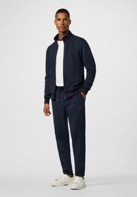Marineblauwe ritsjack met rode accenten, gecombineerd met bijpassende joggingbroek en witte sneakers, met een gladde textuur en een aansluitend ontwerp.