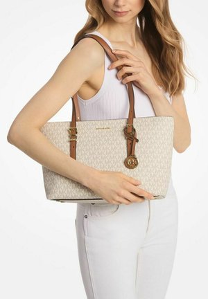 Mujer en top blanco y pantalones sosteniendo un bolso tote beige de Michael Kors con correas marrones y charm de logo en el hombro.