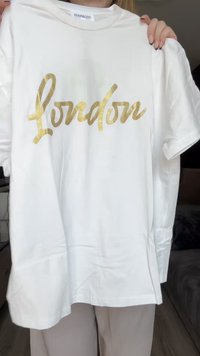 Weißes Baumwoll-T-Shirt mit glänzendem goldenen Schriftzug "London". Kurze Ärmel und runder Ausschnitt. Glatte Textur.