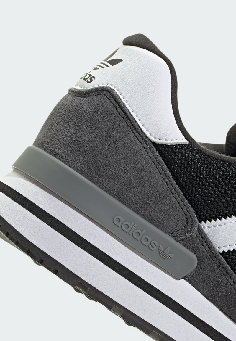 Scarpa da ginnastica nera e grigia con materiali in suede e mesh. Presenta una suola bianca, dettagli grigi e il marchio Adidas ben visibile sul tallone.