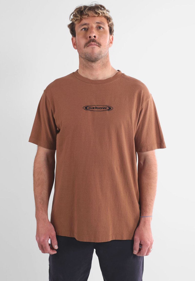 T-shirt en coton marron avec un col rond et des manches courtes, arborant un logo "oxbow" noir au centre. Texture lisse, coupe décontractée.