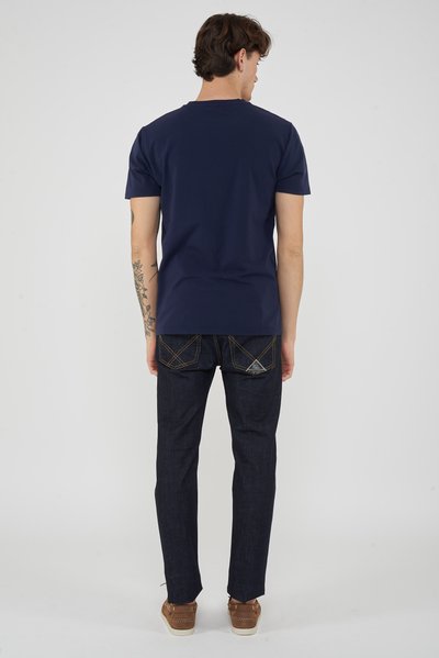 Man with dark hair stands facing away, wearing a navy blue t-shirt, dark jeans with stitched pockets, and brown sneakers.