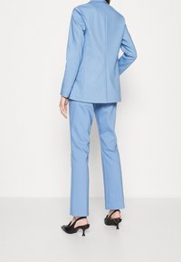 Světle modrý oblek na míru se slim fit sakem a rovnými kalhotami, vyrobený z hladké tkaniny, v kombinaci s černými sandály na podpatcích.