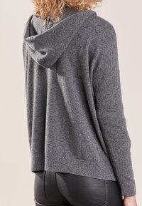Sudadera gris con una textura suave, que presenta una capucha y un corte relajado, vista desde la espalda, combinada con pantalones negros ajustados.
