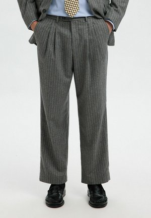 Pantaloni grigi a righe con una vestibilità rilassata, dotati di pieghe frontali e tasche laterali. Abbinati a scarpe di pelle nere.