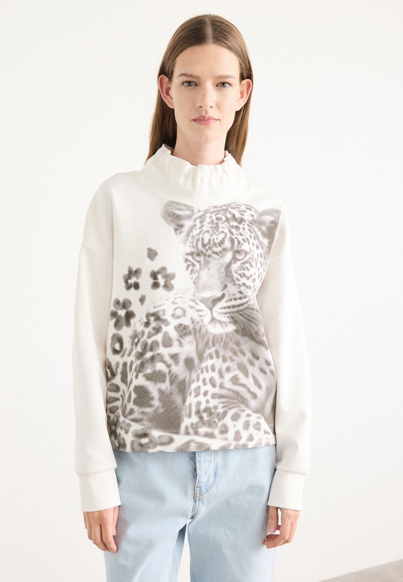 Maglione di cotone con colletto alto a volant, dotato di un motivo leopardato grigio e disegni floreali su uno sfondo crema, abbinato a jeans azzurri chiari.