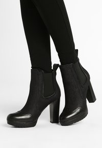 Botas negras hasta el tobillo con un upper de tejido texturizado, paneles laterales elásticos y un tacón grueso. Cuenta con una puntera redondeada y suela de goma.