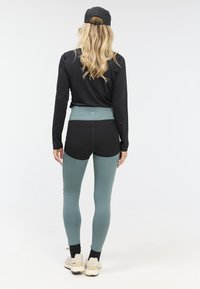 Zwarte lange mouwen top, high-waisted zwarte en groene leggings, strak ontwerp, stoffen textuur zichtbaar, lichte achtergrond, witte sneakers.
