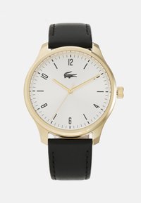 Reloj dorado con una esfera texturada blanca, marcadores de hora negros y correa de cuero negra. Presenta un logotipo a las 3 en punto y una corona.