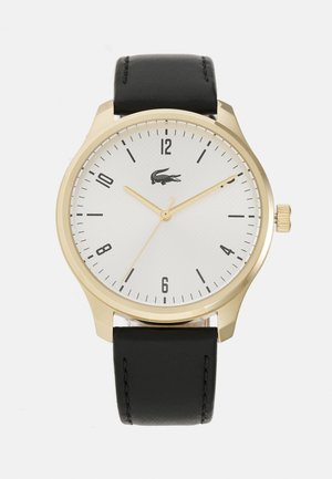 Reloj dorado con una esfera texturada blanca, marcadores de hora negros y correa de cuero negra. Presenta un logotipo a las 3 en punto y una corona.