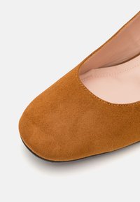 Ballerine en daim marron avec un bout arrondi et un design au cou-de-pied bas. L'intérieur est de couleur claire. La semelle est noire et texturée.