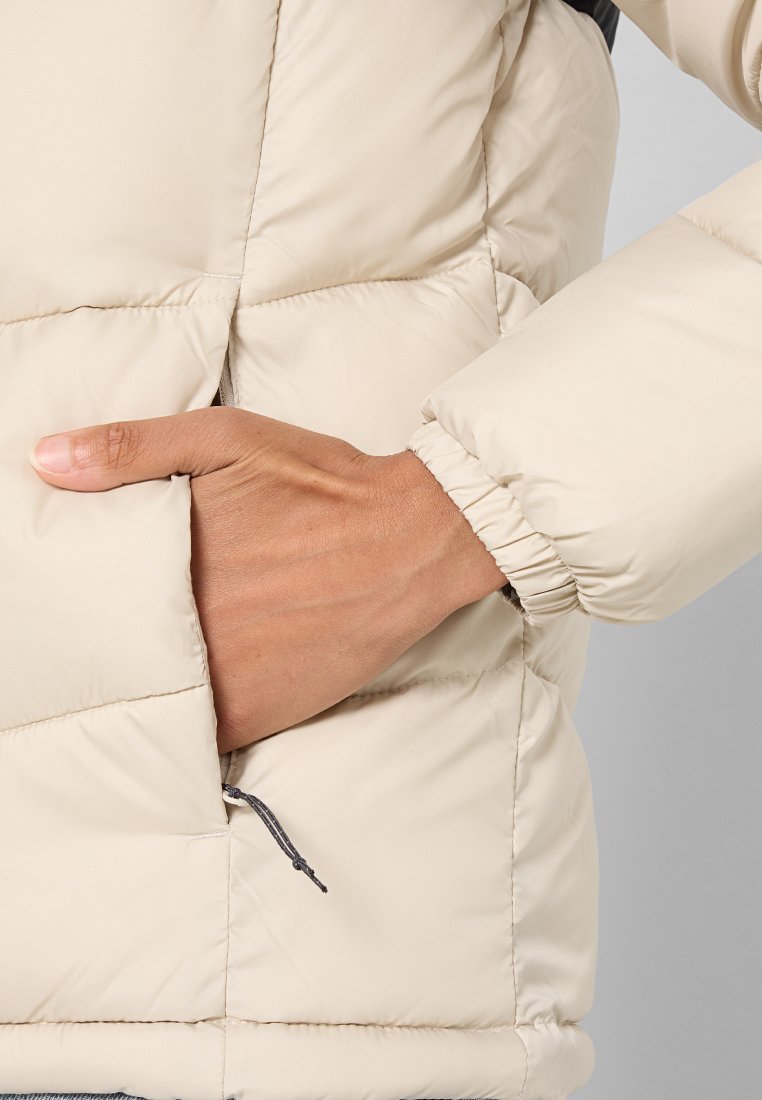 Chaqueta acolchada de color beige claro con un diseño acolchado, puños elásticos y un bolsillo lateral. El material parece suave y aislante.