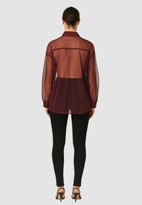 Blusa trasparente color bordeaux con colletto e maniche lunghe, abbinata a leggings neri e tacchi a strappo, che mette in mostra un design a strati.