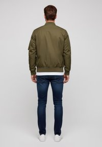 Giacca bomber verde oliva con polsini e orlo a coste, abbinata a jeans blu scuro e scarpe da ginnastica bianche. Tessuto sintetico liscio.