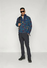 Fleesjakker, mille sinise, punase ja rohelise geomeetrilise mustriga; varustatud lukkude sulgemisega ja kollaste aksessuaaridega; kombineeritud tumedate pükste ja mustade kingadega.