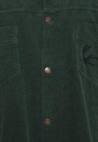 Mörkgrön kordflöjls skjorta med vertikal ribbad textur, guldknappar med tryck och två bröstfickor med broderad logodetalj.