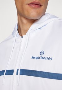 Chaqueta ligera blanca con acentos azules y un logo. Cuenta con cierre de cremallera, capucha y tejido texturizado, adecuada para un uso casual.