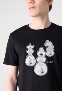 Černé bavlněné tričko s bílou grafikou tří šachových figurek: krále, královny a jezdecka, s nápisem "HUGO" vytištěným bílou barvou.