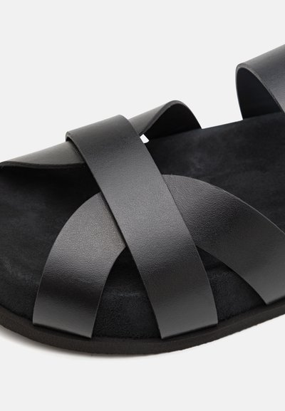Sandale noire avec des brides en cuir croisées, un coussinet de pied en daim souple, des bords arrondis et une semelle plate. La texture paraît lisse et mate.
