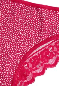 Rode lingerie slip met een wit gespikkeld patroon, voorzien van kanten randafwerking langs de randen voor extra textuur en ontwerp.