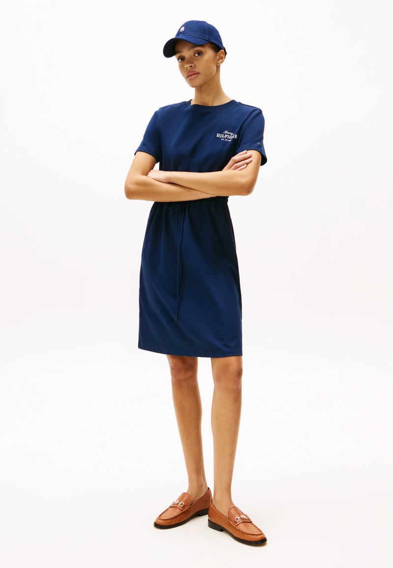 Robe en coton bleu marine à manches courtes avec une taille à cordon de serrage et un accent de logo, associée à des mocassins en cuir marron et une casquette assortie.
