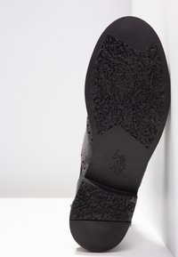 Semelle d’une chaussure habillée noire avec grip texturé, petit logo embossé de joueur de polo à la voûte plantaire, reposant verticalement contre une surface blanche.