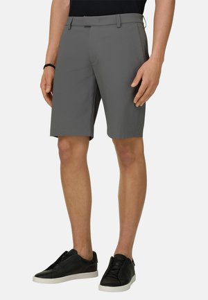 Uomo che indossa pantaloncini grigi su misura, sneakers nere con suola bianca e un braccialetto nero, in piedi davanti a uno sfondo semplice.