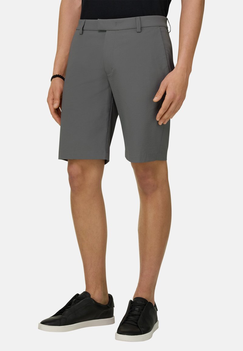 Homme portant un short gris sur mesure, des baskets noires à semelles blanches et un bracelet noir, debout devant un fond uni.