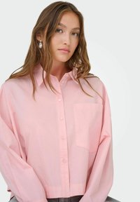 Camicia button-up oversize di un rosa chiaro con maniche lunghe e tasca sul petto. Il tessuto appare liscio con un fit rilassato, indossata in modo casual.