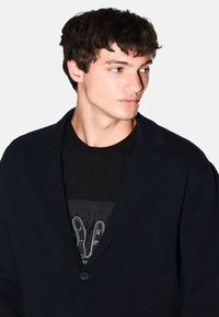 Marineblazer mit glatter Textur, Ein-Knopf-Verschluss und Graslappen, über einem schwarzen T-Shirt mit grafischem Design getragen.