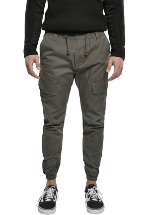 Homme portant un pantalon cargo jogging vert olive avec taille à cordon et des baskets noires avec des lacets blancs, debout sur fond blanc.