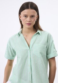 Camisa de manga corta a rayas verdes y blancas con frontales abotonados y un bolsillo en el pecho. Tejido suave con un corte holgado.