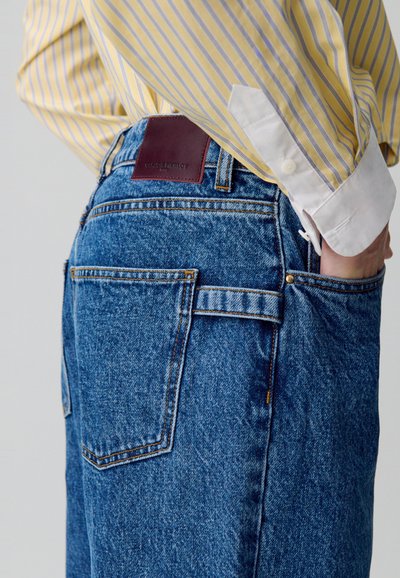 Vaqueros de mezclilla azul con un corte relajado, que cuentan con una etiqueta de marca de cuero marrón, costuras visibles y detalles adicionales en los bolsillos.