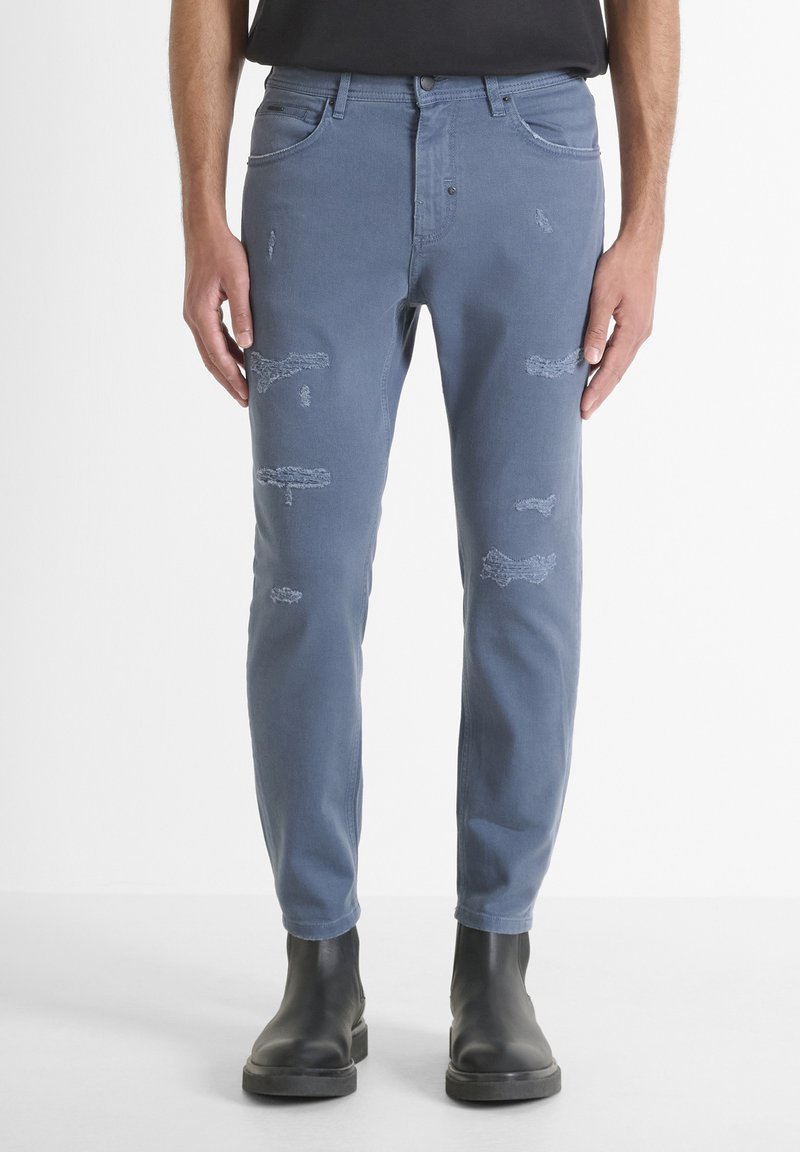 Hombre vistiendo jeans ajustados azules desgastados y botas negras hasta el tobillo, de pie contra un fondo blanco.