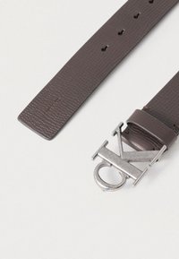 Καφέ ζώνη από δέρμα με υφή, με ασημένια μεταλλική αγκράφα που φέρει τα γράμματα "CK", εμφανίζοντας πολλές τρύπες για ρύθμιση κατά μήκος του λουριού.