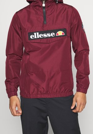 Veste à enfiler bordeaux ellesse avec demi-fermeture éclair et poignets élastiques, portée avec un pantalon noir, montrant une vue partielle des mains et du torse.