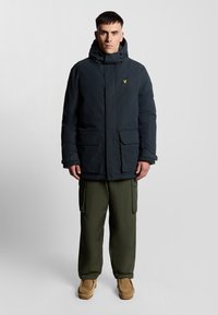 Marinblå pufferjacka med huva, som har två framfickor och en gul logotyp. Kompletterad med olivgröna cargobyxor och beige skor.