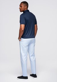 Uomo in polo blu navy, pantaloni azzurri e sneakers nere, in piedi con le mani in tasca, rivolto di lato contro uno sfondo semplice.