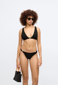 Svart ribbad bikini med djup ringning och guldiga detaljer. Kombinerad med en strukturerad svart väska och runda solglasögon.