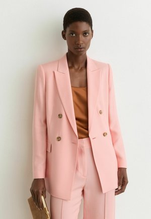 Czarnoskóra kobieta w jasnoróżowej marynarce dwurzędowej i spodniach, brązowej bluzce, trzymająca brązową kopertówkę, stojąca na tle białej ściany.