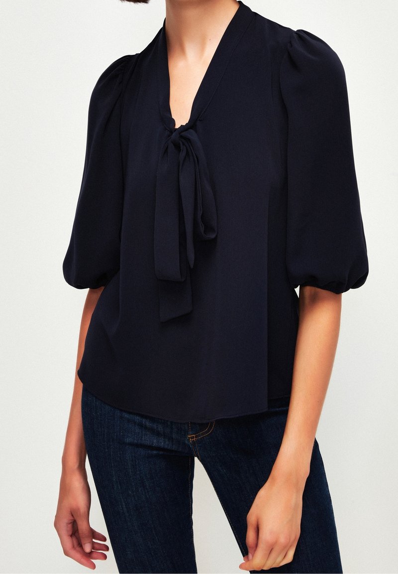 Femme portant un chemisier noir à manches bouffantes mi-longues au coude et un col en V noué, associé à un jean bleu foncé.