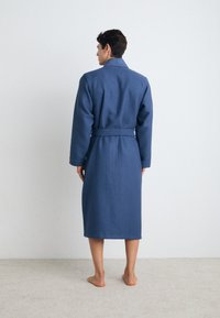 Blå badekåpe med vaffeltekstur, med krage, lange ermer og belte i midjen, som når til midten av leggen, vist på en modell med bare føtter.