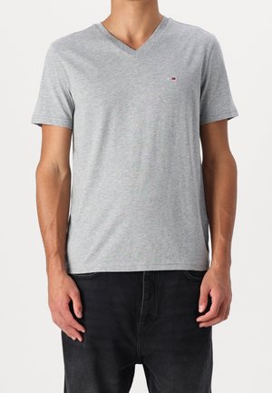 Homme portant un T-shirt à manches courtes en col V gris chiné avec un petit logo rectangulaire sur le côté gauche de la poitrine, associé à un pantalon noir.