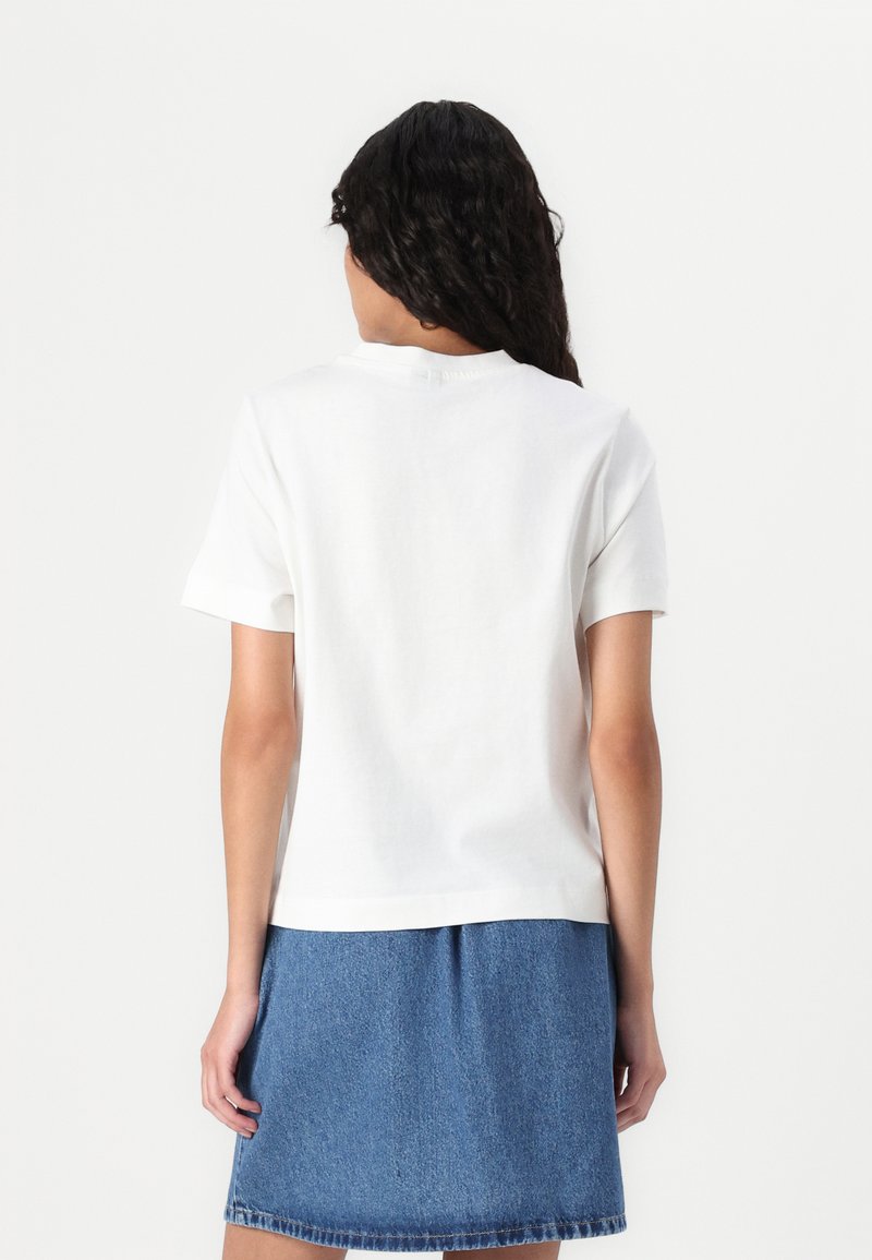 T-shirt blanc à manches courtes avec col rond, en coton, présenté de dos. Associé à une jupe en denim. Texture lisse.