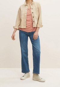 Personne portant une veste beige boutonnée, un haut rose texturé, un jean bleu droit et des baskets montantes beiges, debout devant un fond uni.