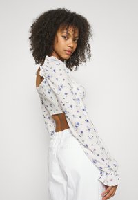 Blusa floral com mangas longas e bufantes, feita de um tecido leve, e com uma parte de trás recortada. Apresenta pequenos padrões de flores azuis e roxas sobre um fundo pálido.