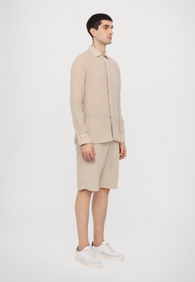 Camisa de linho bege de manga longa com botões e colarinho, acompanhada de calções na mesma tonalidade. Ténis brancos com pormenores em verde completam o visual.