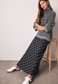 Pull gris flou avec lettres noires, associé à une longue jupe à carreaux noire et grise et des baskets blanches avec des accents noirs.