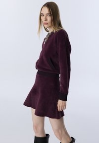 Futrzasty purpurowy sweter z zamkiem i czarnymi ściągaczami, połączony z dopasowaną spódnicą w kształcie dzwonu. Modelka nosi czarne botki.