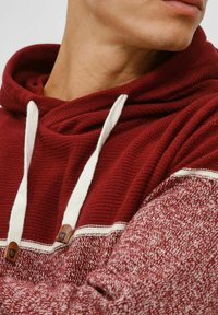 Un hoodie bordeaux avec une section supérieure texturée, des cordons blancs et une quincaillerie en accent marron. Son design dédoublé présente un motif tricot rouge en contraste.